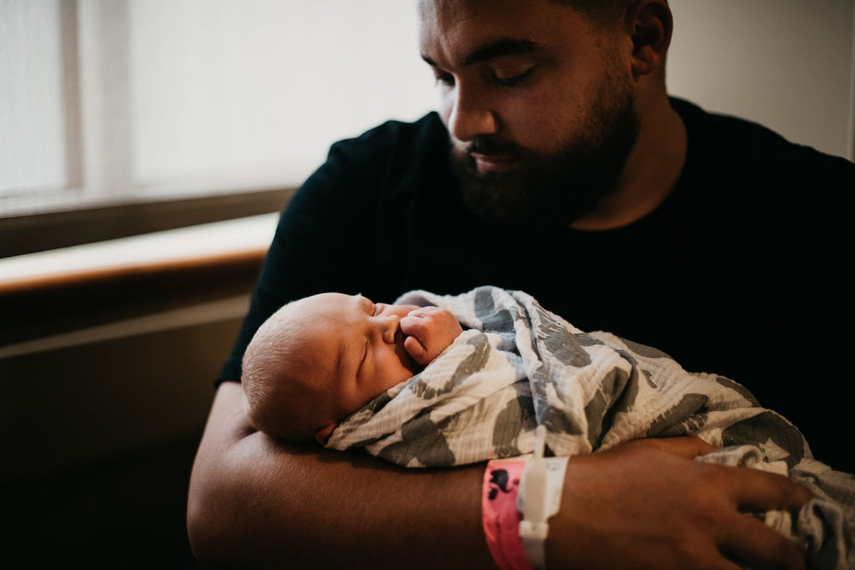 A father holds his sleeping newborn, wrapped snugly in a thin grey patterned muslin