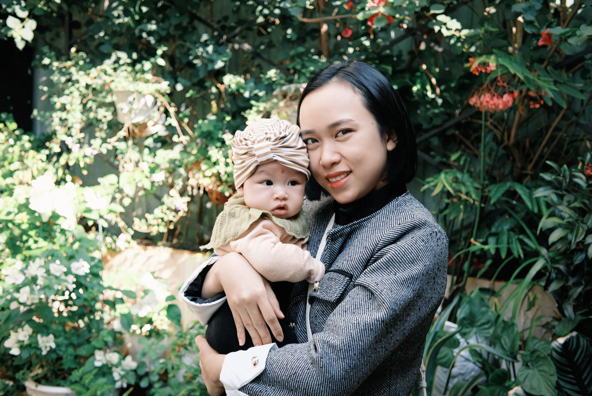 A mother holds her baby in a leafy garden with white flowers behind them