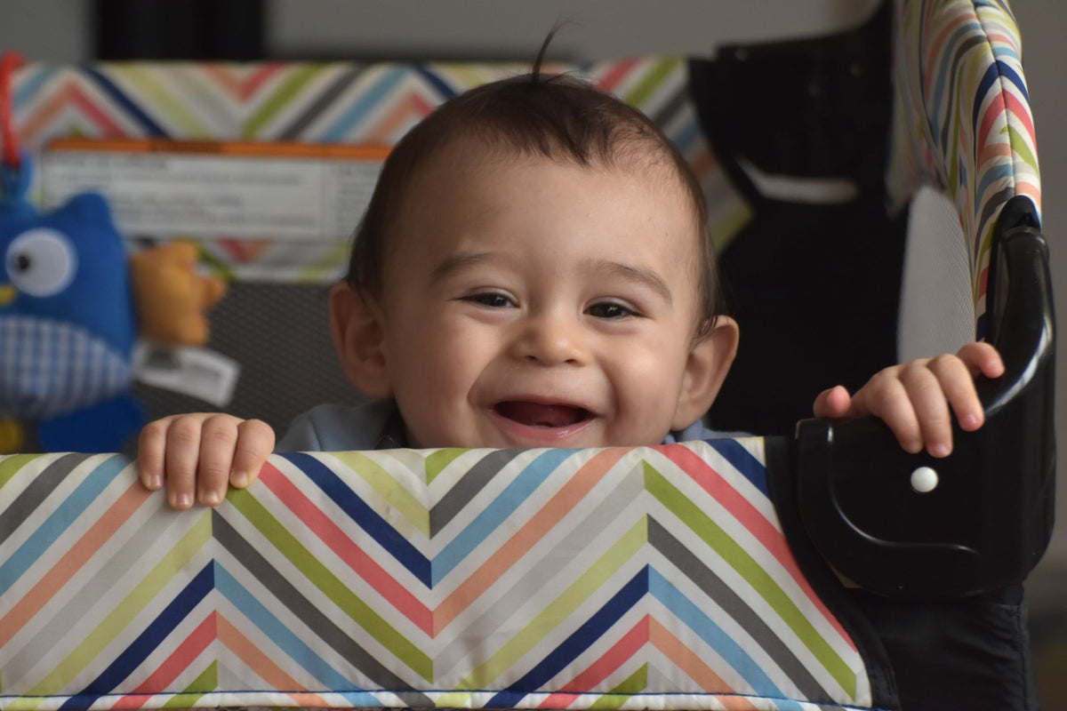 A smiling baby stands safely inside a playpen, holding on to the padded rail