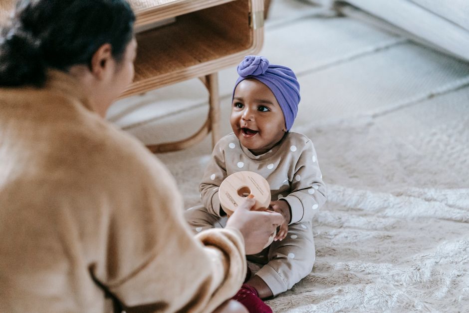 A mother on the floor hands her smiling baby a large wooden rattle