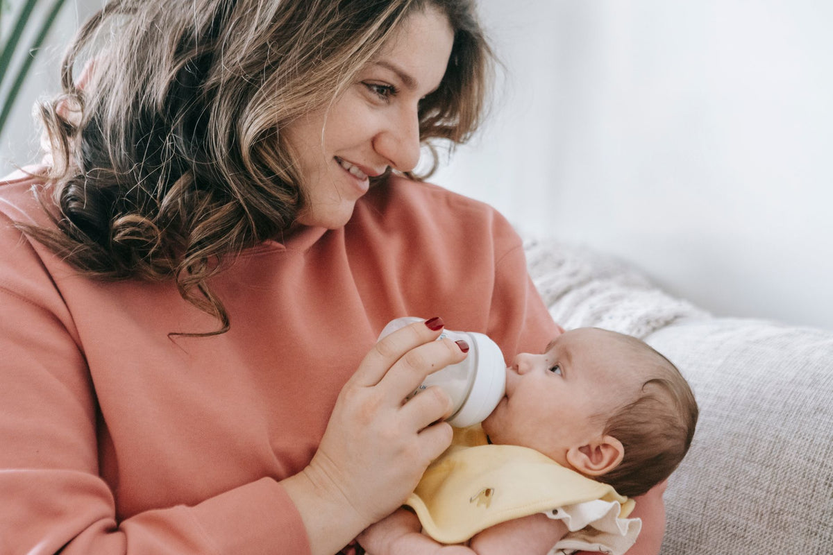A smiling mother bottle-feeds her baby, held close and semi-upright in her arms