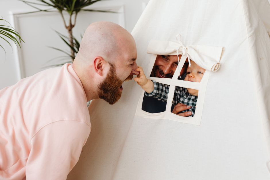 A baby in a play tent reaches through the window to touch a smiling father's nose