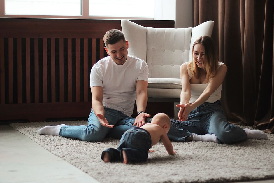 A baby crawls across a rug towards two smiling parents with open arms