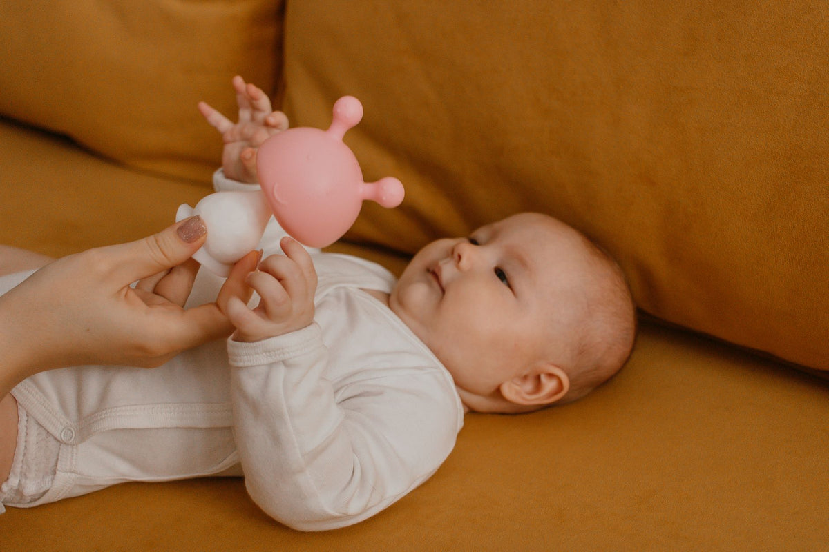 A parent offers a soft silicone teether to a smiling baby lying on their back