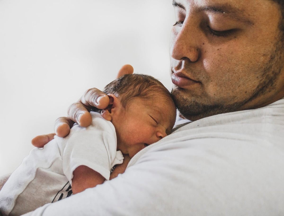 A father holds his sleeping newborn close against his chest in a bright room