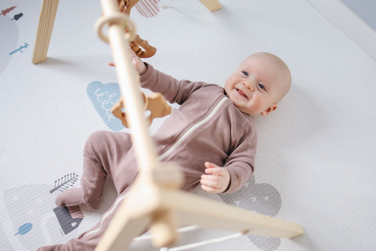 A smiling baby lies on a play mat under a wooden baby gym