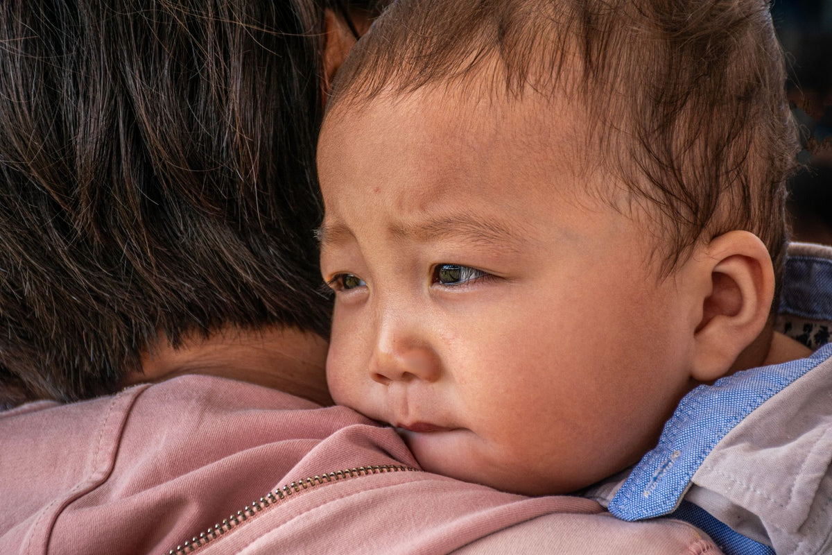 An unsettled baby rests upright on a parent's shoulder, looking tired and teary
