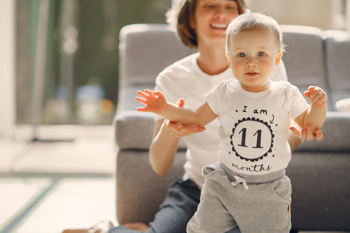 A smiling mother steadies her baby standing on the floor beside a low sofa