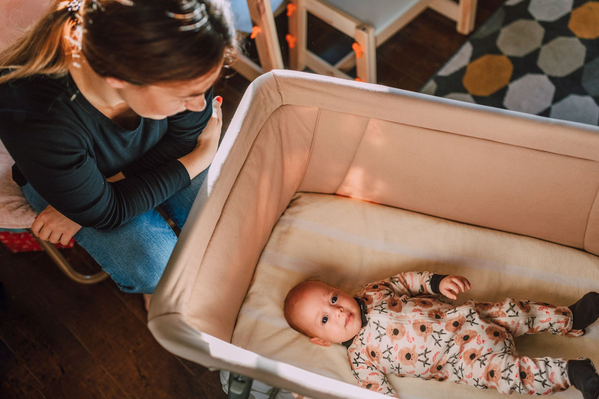 A mother watches her baby lying awake on their back in a clear cot
