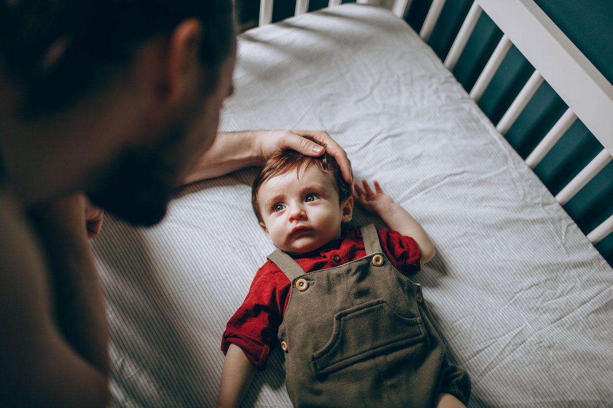 A father gently strokes his baby's head as the baby lies awake in a clear cot