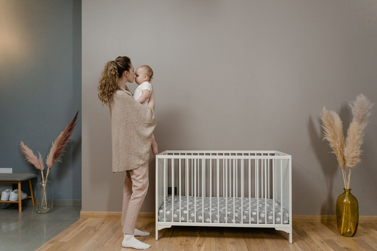 A mother holds her baby beside an empty cot with only a fitted sheet