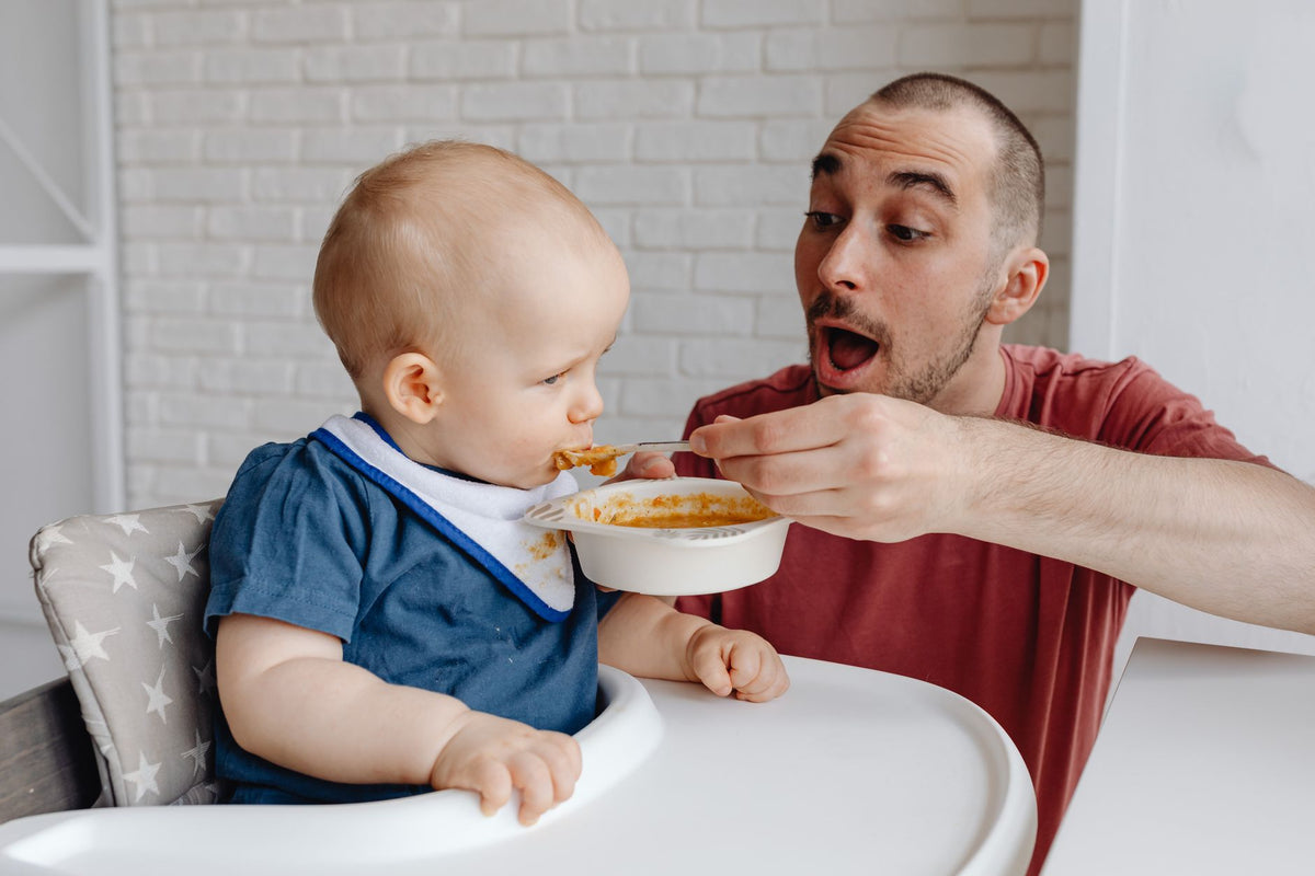 A father spoon-feeds his baby a puree in a high chair at home